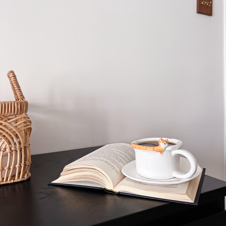 May include: A white coffee mug with a cat figurine on the rim, filled with coffee, sitting on an open book with a brown cover. The mug is on a black surface.