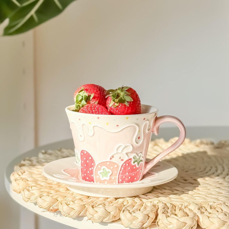 Op de afbeelding: Een roze keramische theekop met een aardbeienontwerp en een wit schoteltje. De theekop is gevuld met verse aardbeien.