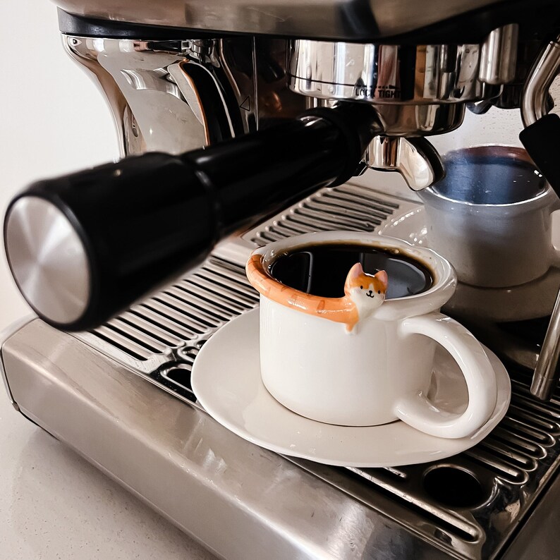 May include: A white ceramic coffee mug with a cat design and a brown handle. The mug is filled with coffee and is sitting on a white saucer. The mug is in front of a silver espresso machine.
