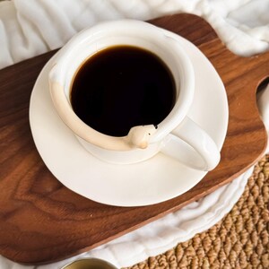 May include: A white ceramic coffee cup and saucer set filled with dark coffee. The cup features a unique design with a bear-shaped handle. The set is placed on a wooden serving tray, creating a cozy and inviting aesthetic.