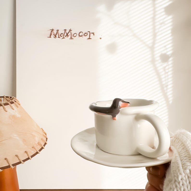 May include: A white ceramic mug with a dachshund dog figurine on the rim, resting on a matching saucer. The dog is black and brown with a pink nose. The mug is held by a person wearing a white sweater.