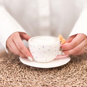 May include: A white ceramic coffee mug with a colorful confetti pattern and a small cat figurine on the handle. The mug is filled with a light brown liquid and is sitting on a white saucer.