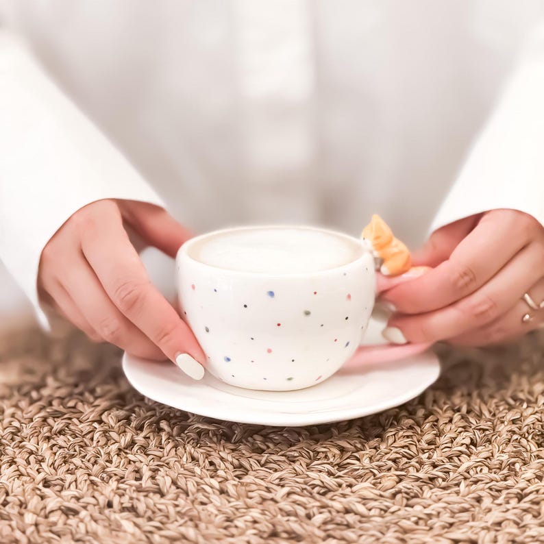 May include: A white ceramic coffee mug with a colorful confetti pattern and a small cat figurine on the handle. The mug is filled with a light brown liquid and is sitting on a white saucer.