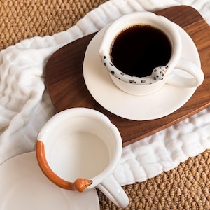 May include: Two white ceramic coffee cups with dog-shaped handles. One cup is filled with dark coffee and sits on a saucer. The other cup is empty. The handles are designed to look like dachshunds, with one featuring black spots and the other a brown and orange color scheme.
