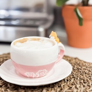 May include: A pink and white ceramic coffee mug with a cute orange cat figurine on the rim. The mug is filled with coffee and is sitting on a white saucer.