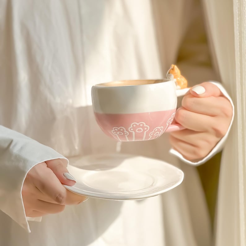 May include: A pink and white ceramic mug with a cat paw print design. The mug has a small cat figurine on the handle. The mug is filled with a light brown liquid.
