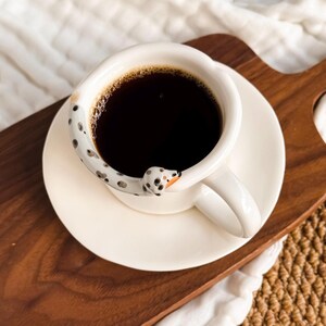 May include: A white ceramic coffee cup and saucer set filled with dark coffee. The handle is shaped like a Dalmatian dog with black spots and an orange nose. The cup is on a wooden tray.