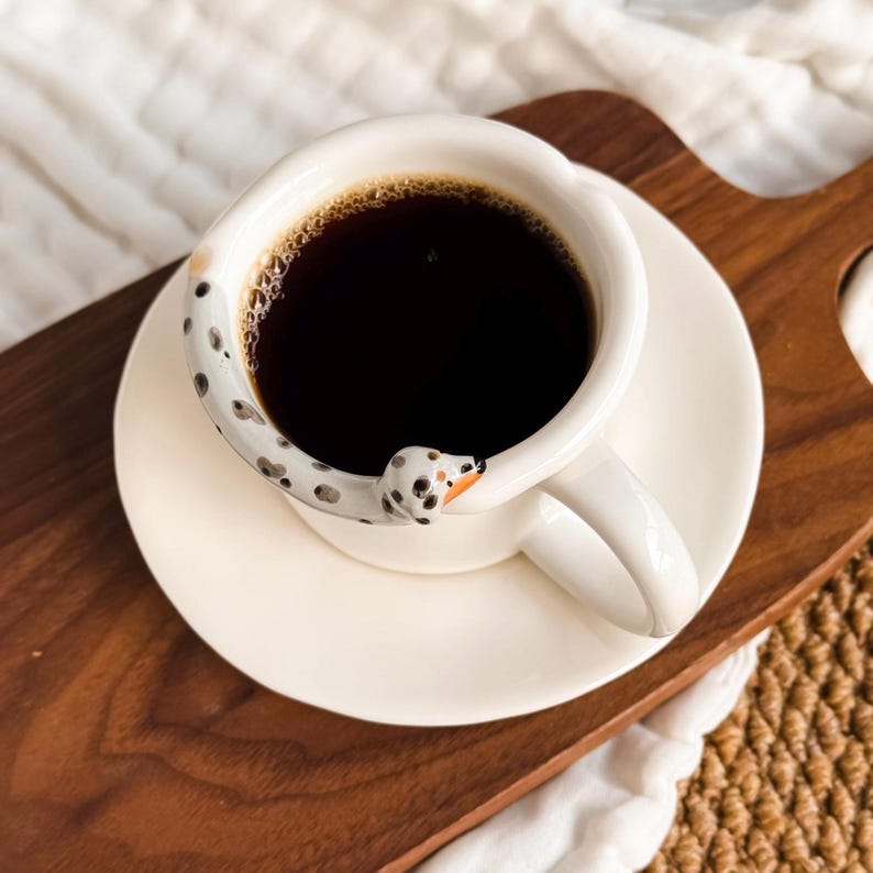 May include: A white ceramic coffee cup and saucer set filled with dark coffee. The handle is shaped like a Dalmatian dog with black spots and an orange nose. The cup is on a wooden tray.