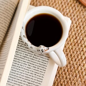 May include: A white ceramic coffee mug filled with dark coffee, featuring a Dalmatian dog design on the handle. The mug rests on an open book with printed text, suggesting a cozy reading setting.