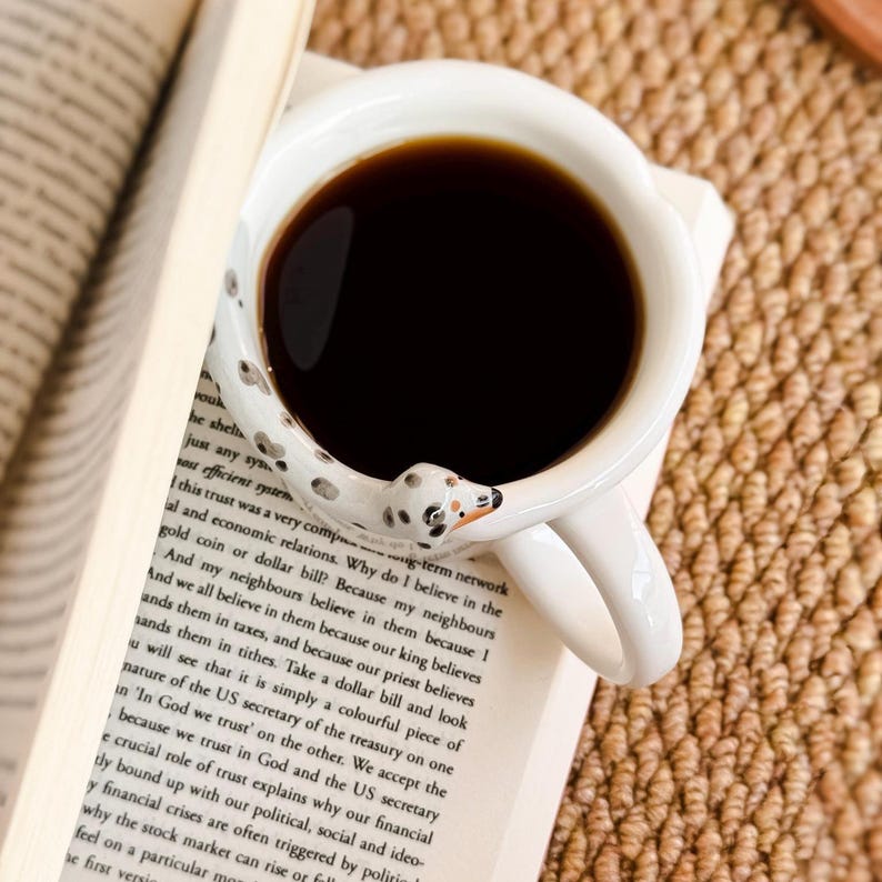 May include: A white ceramic coffee mug filled with dark coffee, featuring a Dalmatian dog design on the handle. The mug rests on an open book with printed text, suggesting a cozy reading setting.