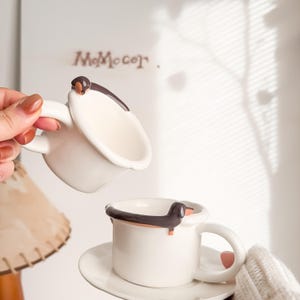 May include: White ceramic mugs with a dachshund dog design. The mugs have a brown and black dachshund dog accent on the rim and handle. One mug is on a saucer. The background has the text "MoMoCor".