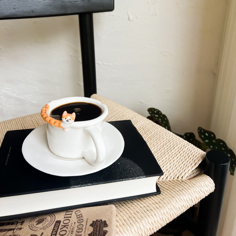 May include: A white ceramic coffee mug with a brown cat figurine peeking out of the top. The mug is filled with coffee and is sitting on a black book with a white saucer underneath. The book is on a woven chair.