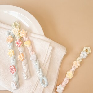 May include: Three pastel-colored, twisted, candy cane-like sticks with various pastel-colored flower and heart shapes. The sticks are on a white surface.