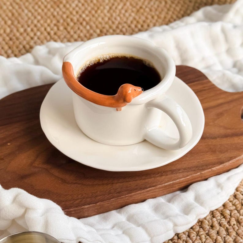 May include: A white ceramic coffee mug filled with dark coffee, resting on a matching saucer. A brown dachshund dog decorates the rim of the mug. The mug and saucer are on a wooden tray.