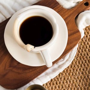 May include: A white ceramic coffee cup filled with dark liquid, resting on a matching saucer. The handle is designed as a polar bear, with the bear's head extending over the cup's rim. The cup sits on a wooden cutting board.
