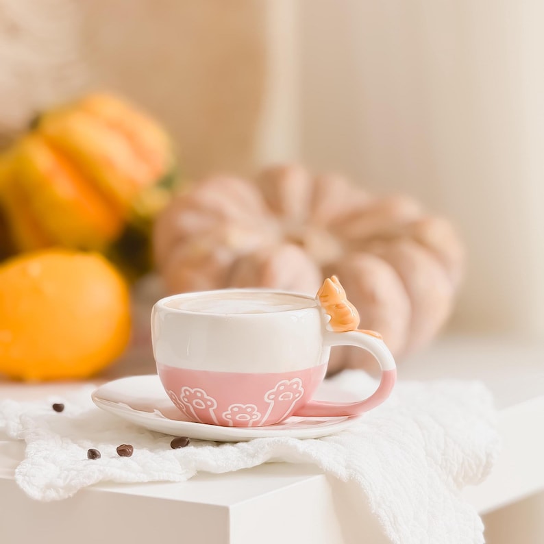 May include: A pink and white ceramic coffee mug with a cat figurine on the handle. The mug is sitting on a white saucer and a white linen napkin. There are pumpkins in the background.