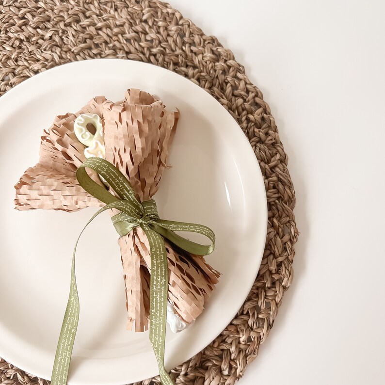 May include: A white plate with a brown paper-wrapped bouquet of white and green candies tied with a green ribbon. The plate is on a woven brown placemat.