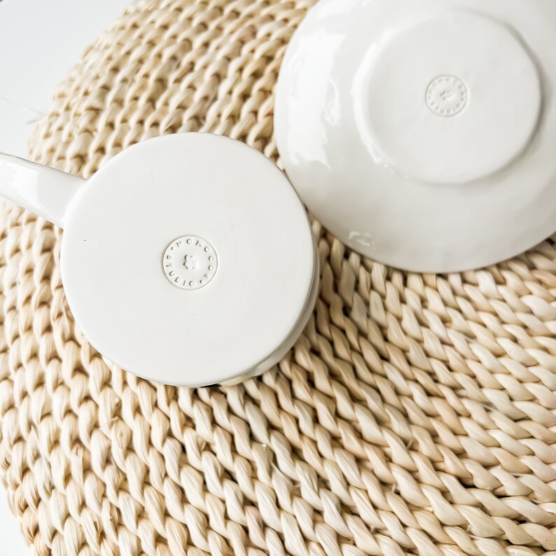 May include: White ceramic teacup and saucer with a gold logo that reads "FULIC" in a circle. The cup and saucer are sitting on a woven straw mat.