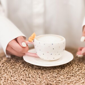 May include: A white ceramic coffee cup with pink accents and a cat figurine on the handle. The cup is filled with coffee and is sitting on a white saucer. The saucer is on a woven brown surface.
