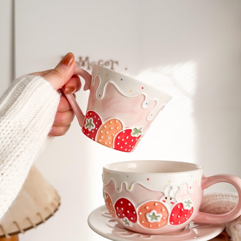 Op de afbeelding: Twee roze keramische mokken met een aardbeienontwerp. De mokken hebben een witte glazuur met een roze en witte marmereffect. De mokken zijn versierd met een aardbeienontwerp en een witte glazuur die eruitziet als gesmolten glazuur. E&eacute;n mok wordt vastgehouden door een hand, terwijl de andere mok op een schotel staat.
