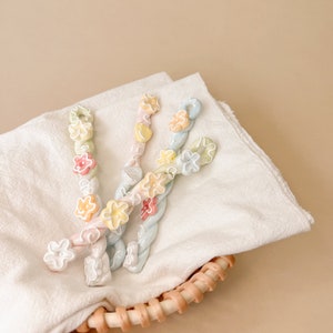 May include: Three pastel-colored hair clips with flower accents. The clips are twisted and have a floral design. The clips are on a white fabric background.