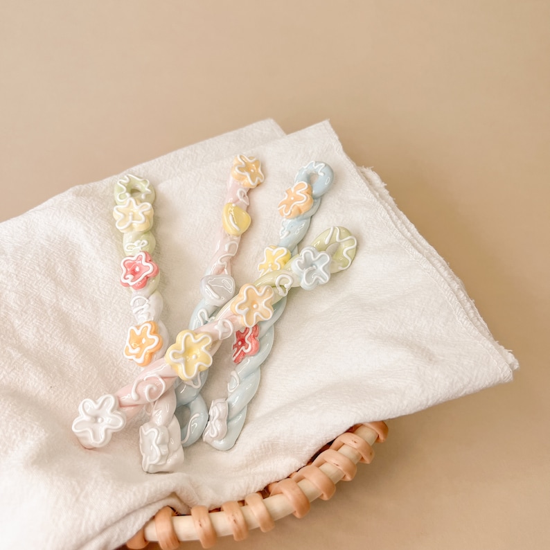 May include: Three pastel-colored hair clips with flower accents. The clips are twisted and have a floral design. The clips are on a white fabric background.