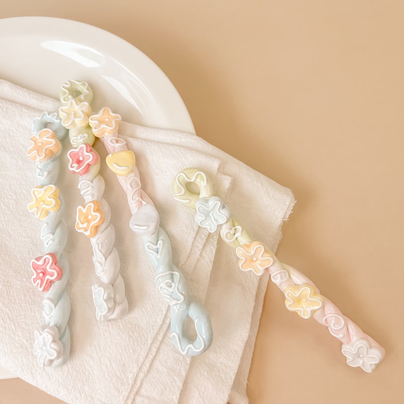 May include: Four pastel-colored twisted ceramic hair clips with flower-shaped embellishments. The clips are arranged on a white surface.