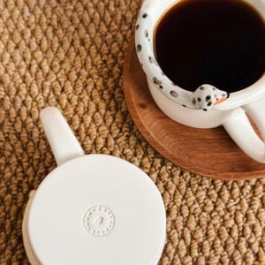 May include: A white ceramic mug filled with dark liquid, featuring a Dalmatian dog design on the handle and rim. The mug sits on a wooden coaster, next to a white ceramic lid. The mug is on a woven brown surface.