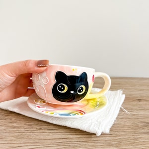 Può includere: Una tazza da tè in ceramica rosa e gialla con un design a forma di gatto nero. La tazza ha un piattino bianco con un design a forma di arcobaleno.