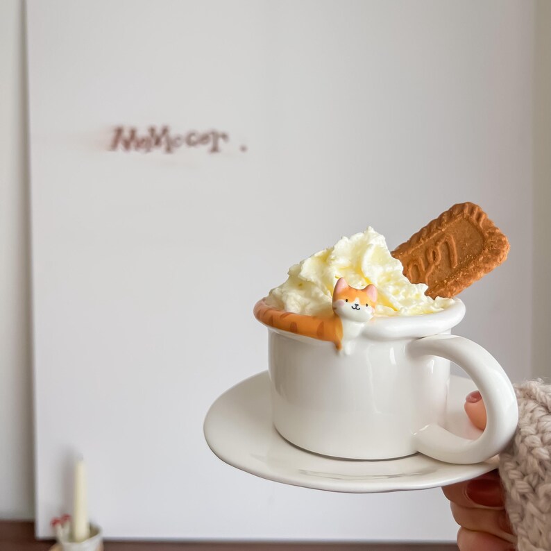 May include: A white ceramic mug with a cat figurine peeking out of the whipped cream on top. A biscuit is resting on the rim of the mug. The mug is on a white saucer.