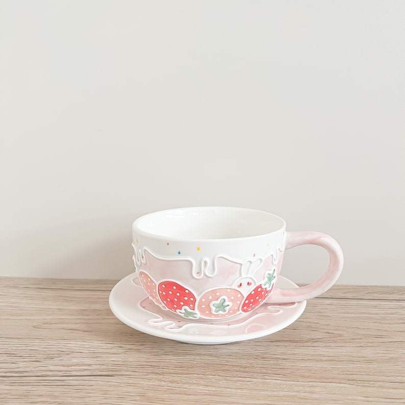 Op de afbeelding: Een roze theekopje en schotel met een witte rand en een aardbeidesontwerp. Het kopje heeft een wit handvat en een roze glazuur met witte gedruppelde glazuur en aardbeien.