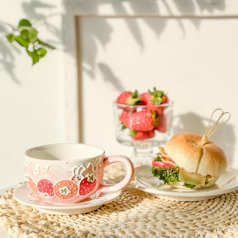 Op de afbeelding: Een roze keramische theekop met een aardbei- en konijnenontwerp, een bijpassend schoteltje en een bord met een sandwich op een geweven placemat.