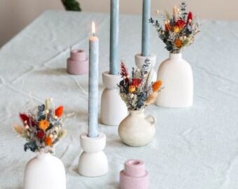 May include: A table setting with three blue candles in white holders, two small white vases with dried flowers, and two pink candle holders.