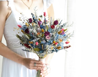 May include: A bouquet of dried flowers in shades of blue, purple, pink, red, yellow, and orange. The bouquet is tied with a white ribbon and includes dried grasses and other foliage.