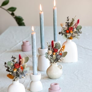 Puede incluir: Una mesa decorada con tres velas azules en candelabros blancos, tres pequeños jarrones blancos con flores secas y dos candelabros rosas.