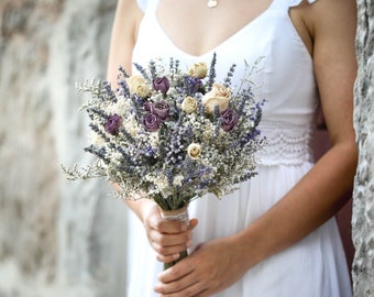 May include: A bride holds a bouquet of dried lavender, white, and purple flowers. The bouquet is tied with a white ribbon.