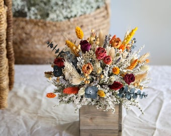 Könnte beinhalten: Eine Holzkiste gefüllt mit einem Trockenblumenarrangement. Das Arrangement besteht aus einer Vielzahl von Trockenblumen in Rot-, Orange-, Gelb- und Weißtönen. Die Blumen sind in einem rustikalen Stil angeordnet, wobei einige Stiele aus der Kiste herausragen.