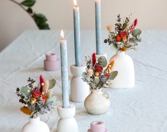 May include: A table setting with three blue candles in white candle holders, three small white vases with dried flowers, and two pink candle holders.