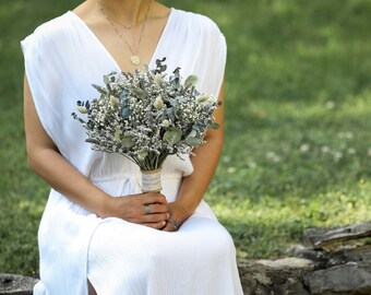 May include: A person in a white dress holds a bouquet of dried flowers. The bouquet is made up of white and green flowers and greenery.