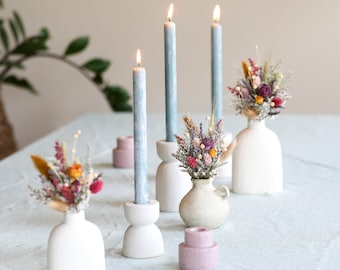 May include: A table setting with three blue candles in white holders, each with a small vase of dried flowers. The flowers are a mix of white, pink, purple, and orange.