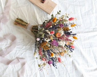 Könnte beinhalten: Ein Trockenblumenstrauß mit verschiedenen Farben wie Rosa, Lila, Orange und Gelb. Der Strauß ist mit Bindfaden und Spitze gebunden und liegt auf einer weißen Oberfläche. Ein hellrosa Band ist in der Nähe drapiert.