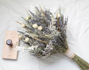 May include: A dried flower bouquet featuring lavender, white roses, and various dried grasses. The bouquet is tied with twine and lace. A wooden stamp with a dark handle rests nearby. The arrangement is set against a white background.