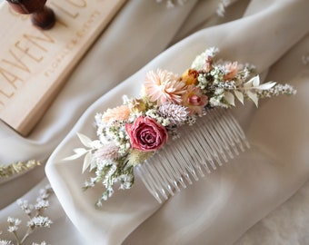 May include: A white plastic hair comb with dried flowers attached. The flowers are pink, peach, and white. The comb is on a white fabric background.