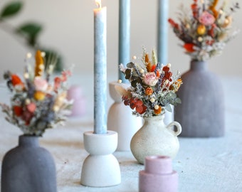 May include: A table setting with three small vases filled with dried flowers and a candle in a white holder. The vases are all different shapes and colors, and the flowers are a mix of orange, pink, and white.