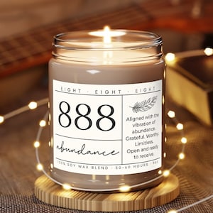 May include: A brown glass candle with a white label that reads "EIGHT EIGHT EIGHT 888 abundance" and "Aligned with the vibration of abundance. Grateful. Worthy. Limitless. Open and ready to receive. 100% Soy Wax Blend 50-60 Hours".