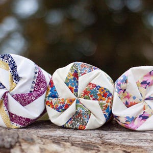 May include: Three cylindrical fabric rolls featuring a pinwheel-style spiral pattern. Each roll displays a unique combination of white fabric segments alternating with colourful floral or paisley prints in shades of blue, yellow, red, and pink.