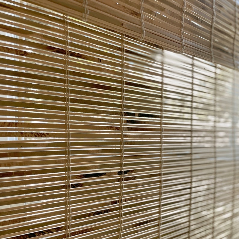 May include: Close-up of a natural woven bamboo window blind in a light brown colour. The blind is made of thin strips of bamboo that are woven together with white string.