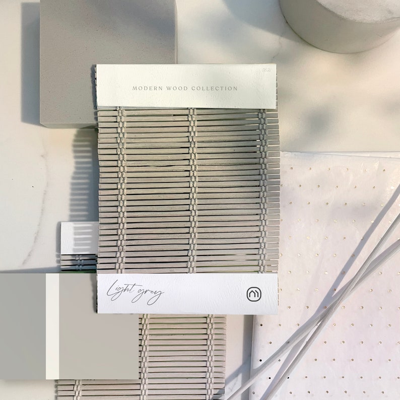 May include: A sample of light grey wood slat blinds from the Modern Wood Collection. The sample is labelled "light grey" and has a small logo in the corner.