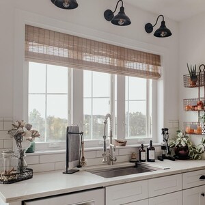 May include: A white kitchen with a farmhouse sink, white cabinets, and a window with a brown roman blind. There are three black pendant lights above the sink.