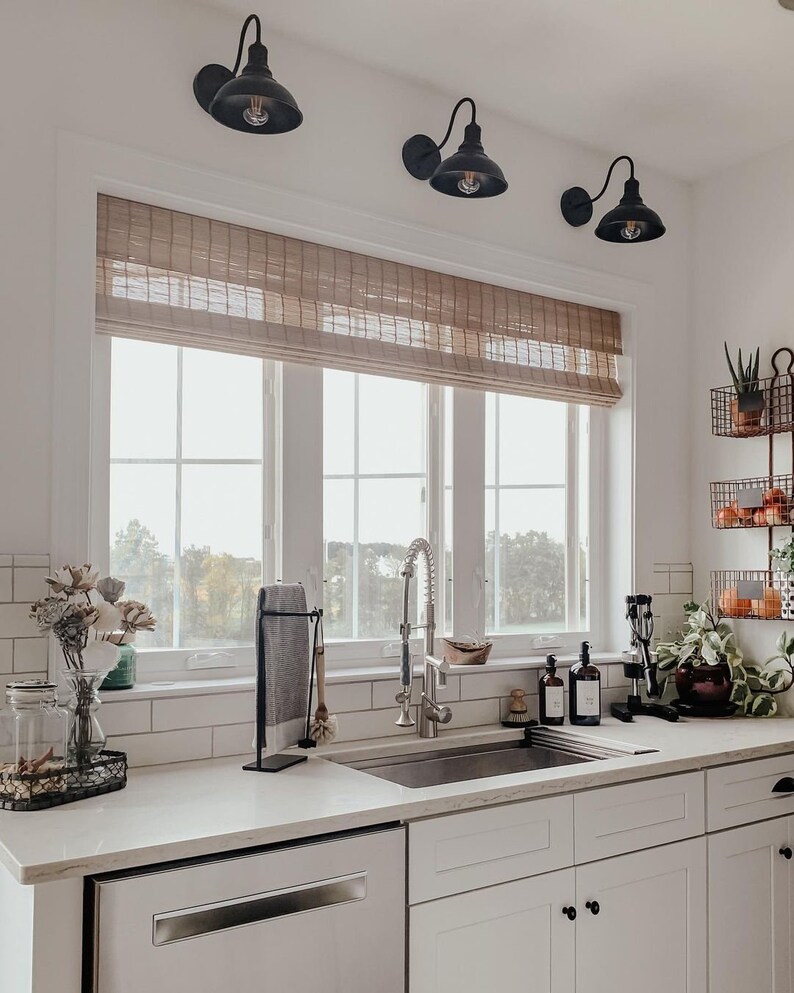 May include: A white kitchen with a farmhouse sink, white cabinets, and a window with a brown roman blind. There are three black pendant lights above the sink.